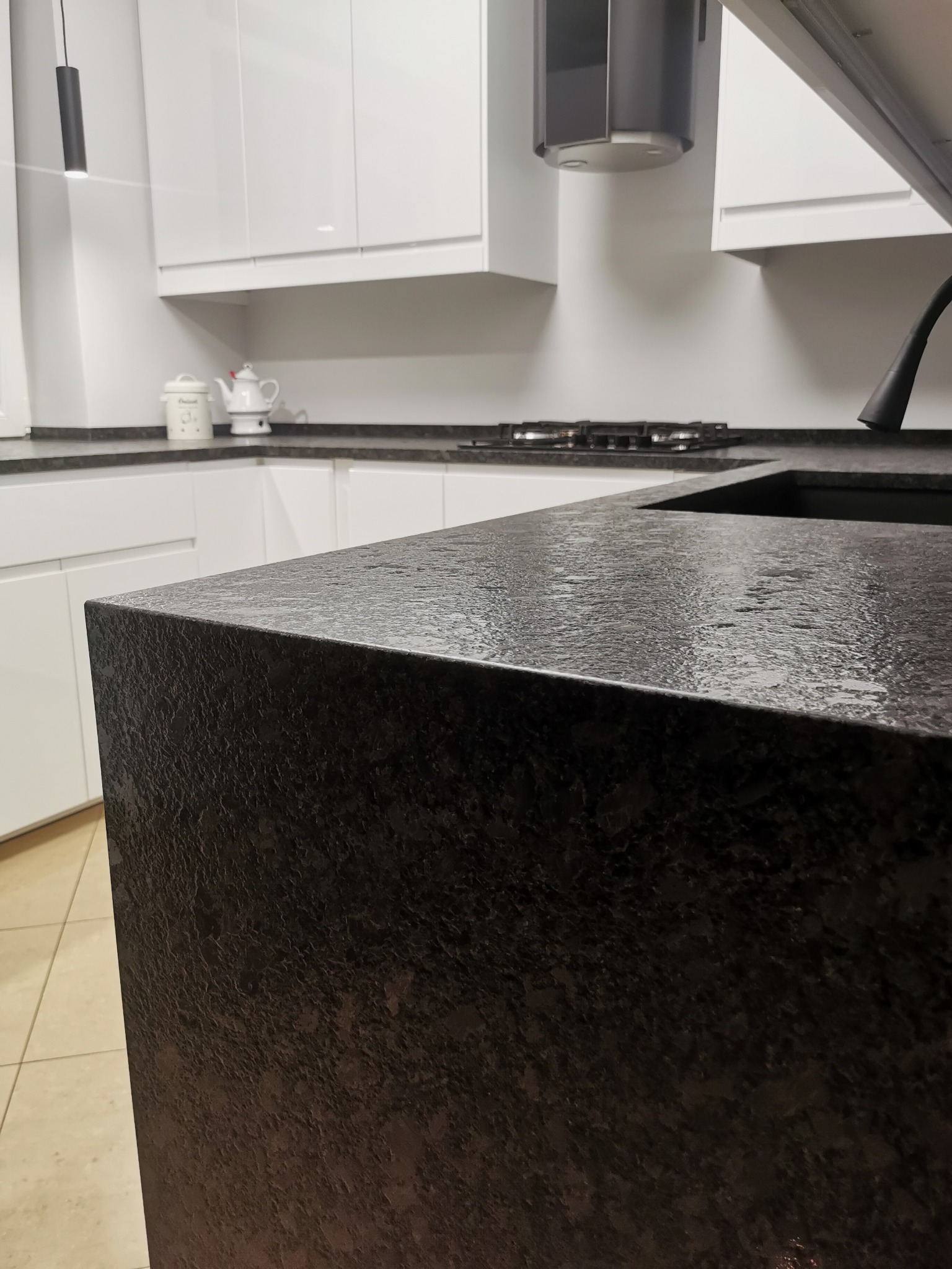 Czarny, teksturowany blat kamienny w nowoczesnej kuchni z białymi szafkami, widoczne krople wody na powierzchni, oświetlenie punktowe