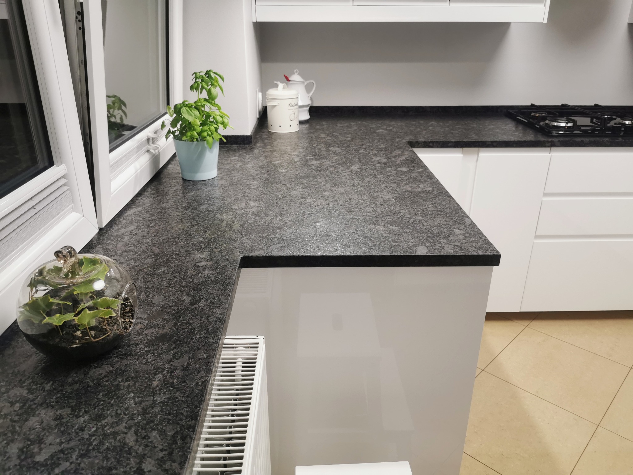 Szary blat kuchenny z ciemnymi plamkami, z roślinami w doniczce i szklanej kuli, białe szafki kuchenne i okno w tle.