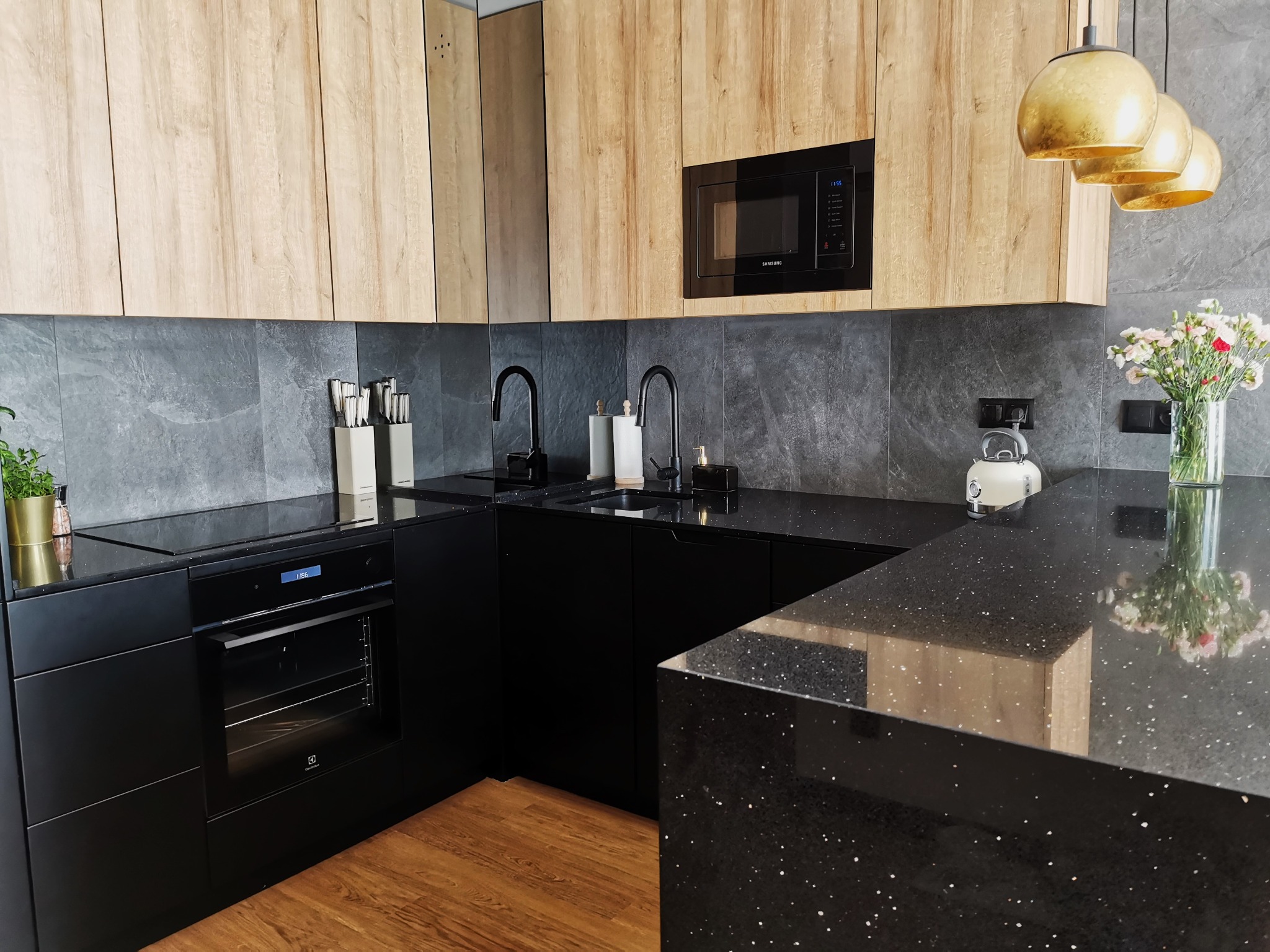 Nowoczesna kuchnia z czarnymi szafkami, drewnianymi górnymi szafkami i czarnym blatem z drobinkami, oświetlona złotymi lampami wiszącymi.