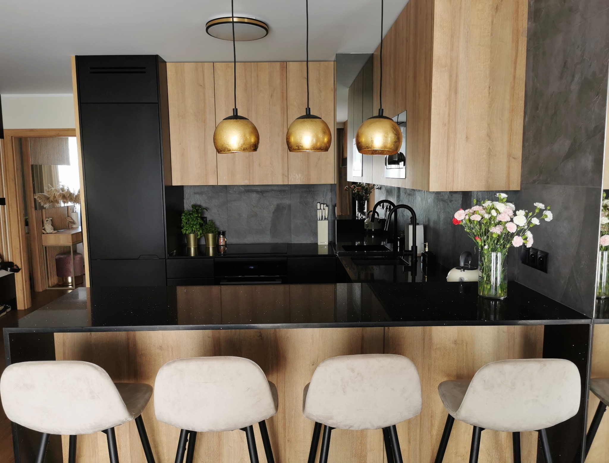 Nowoczesna kuchnia z czarnym blatem o wysokim połysku, drewnianymi szafkami, trzema złotymi lampami wiszącymi i czterema krzesłami barowymi z beżową tapicerką.