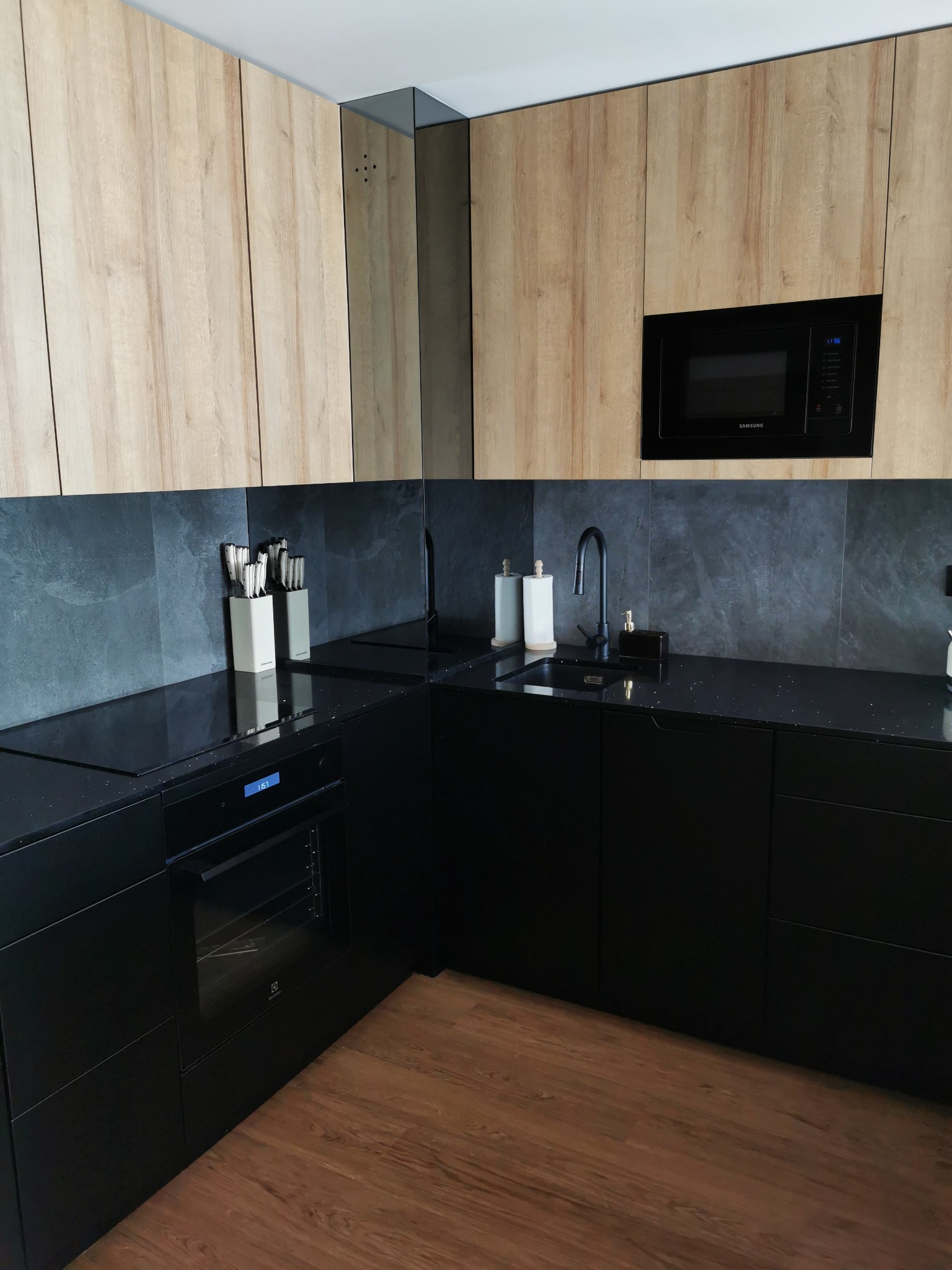 Czarna, błyszcząca powierzchnia blatu kuchennego z widocznymi drobinkami, komponująca się z ciemnoszarą ścianą i czarnymi szafkami, uzupełniona jasnymi, drewnopodobnymi frontami szafek górnych...