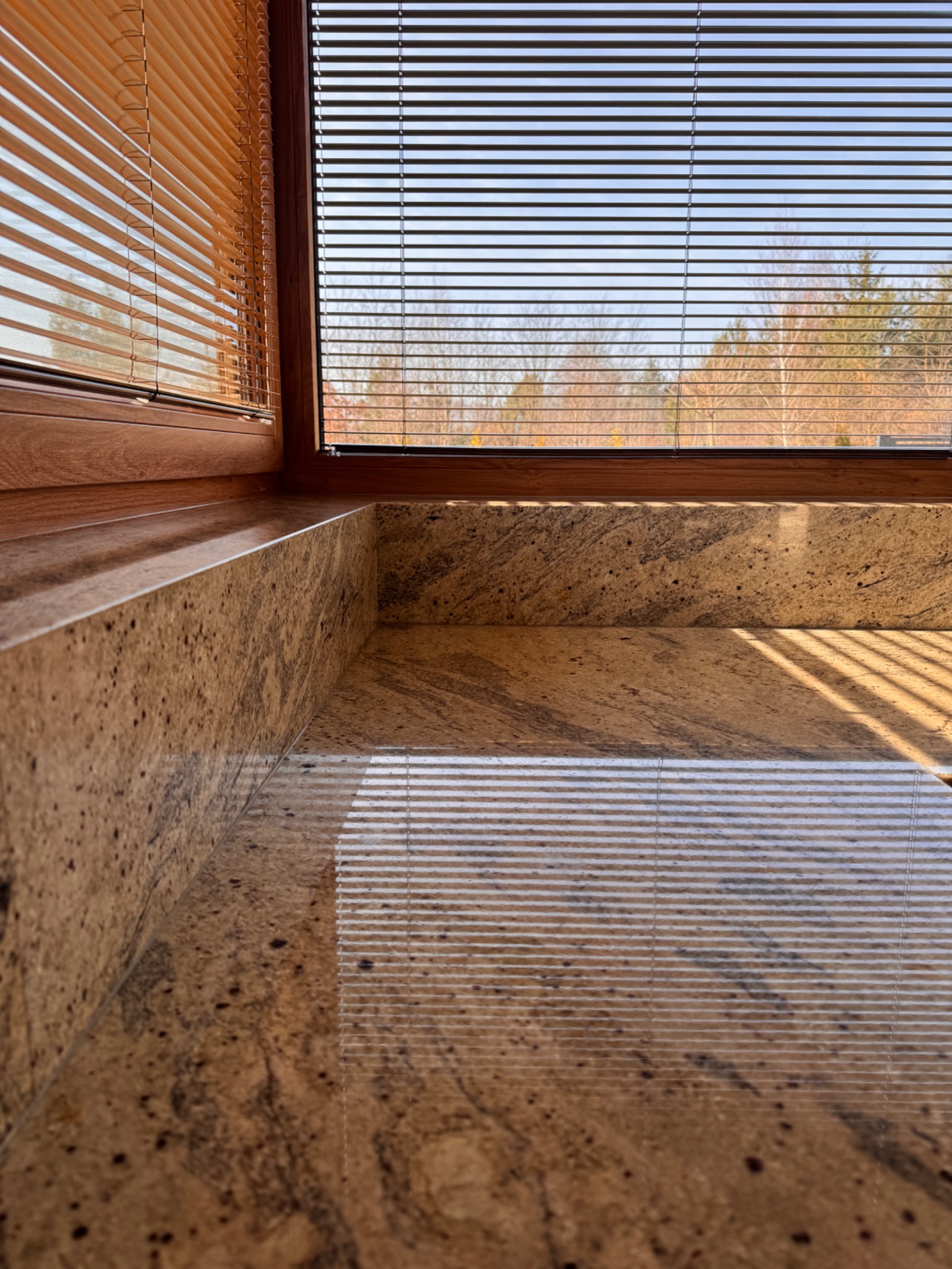 Beżowy parapet z kamienia naturalnego wnęki okiennej, z widocznymi żaluzjami i fragmentem krajobrazu za oknem. Polerowana powierzchnia odbija światło.