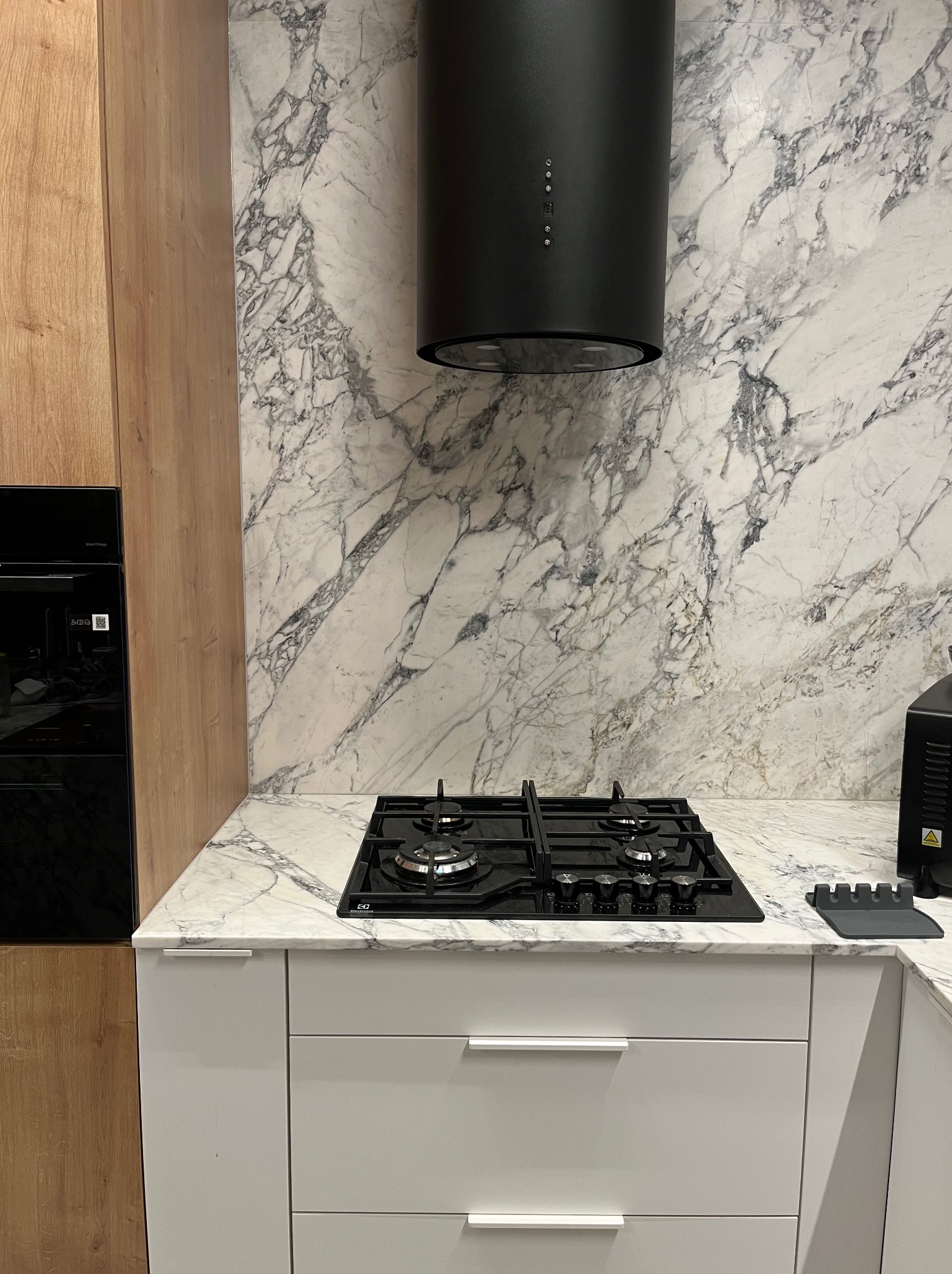 Biały blat kuchenny z marmurowym wzorem, czarna płyta gazowa i okap. Jasne drewniane szafki po lewej stronie. Minimalistyczny design.