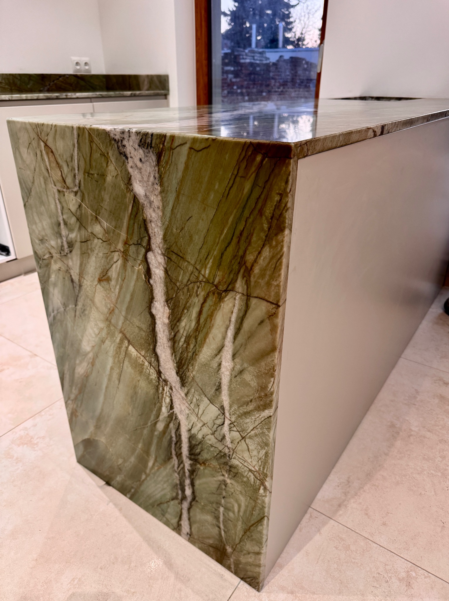 Zielony blat kamienny z wyraźnym żyłkowaniem, zamontowany na wyspie kuchennej. Widoczne polerowane wykończenie i beżowa podłoga z płytek.