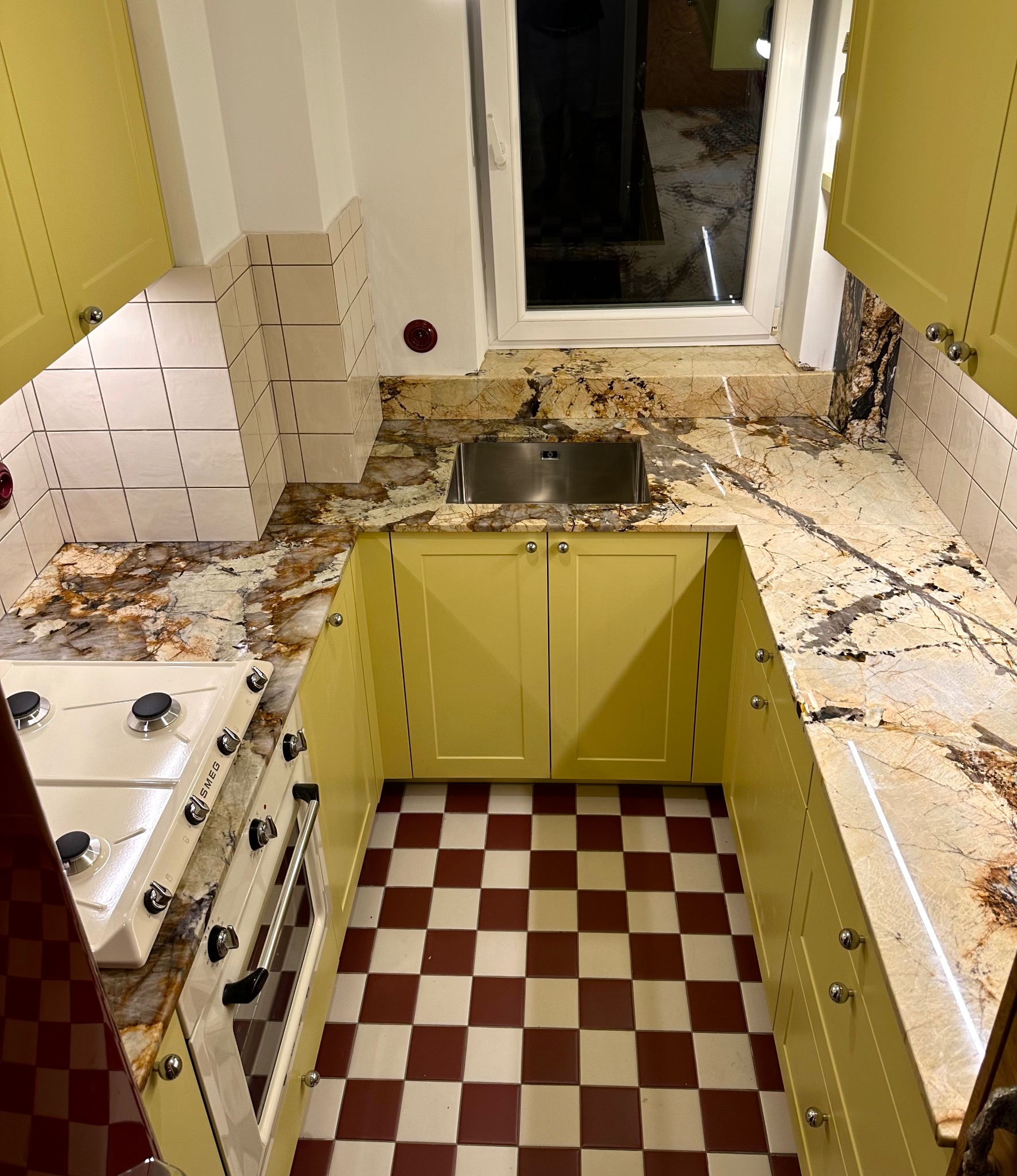 Kuchnia w stylu retro z blatem kamiennym o unikalnym wzorze, żółtymi szafkami i biało-bordową podłogą w szachownicę. Widoczny zlew i fragment kuchenki.