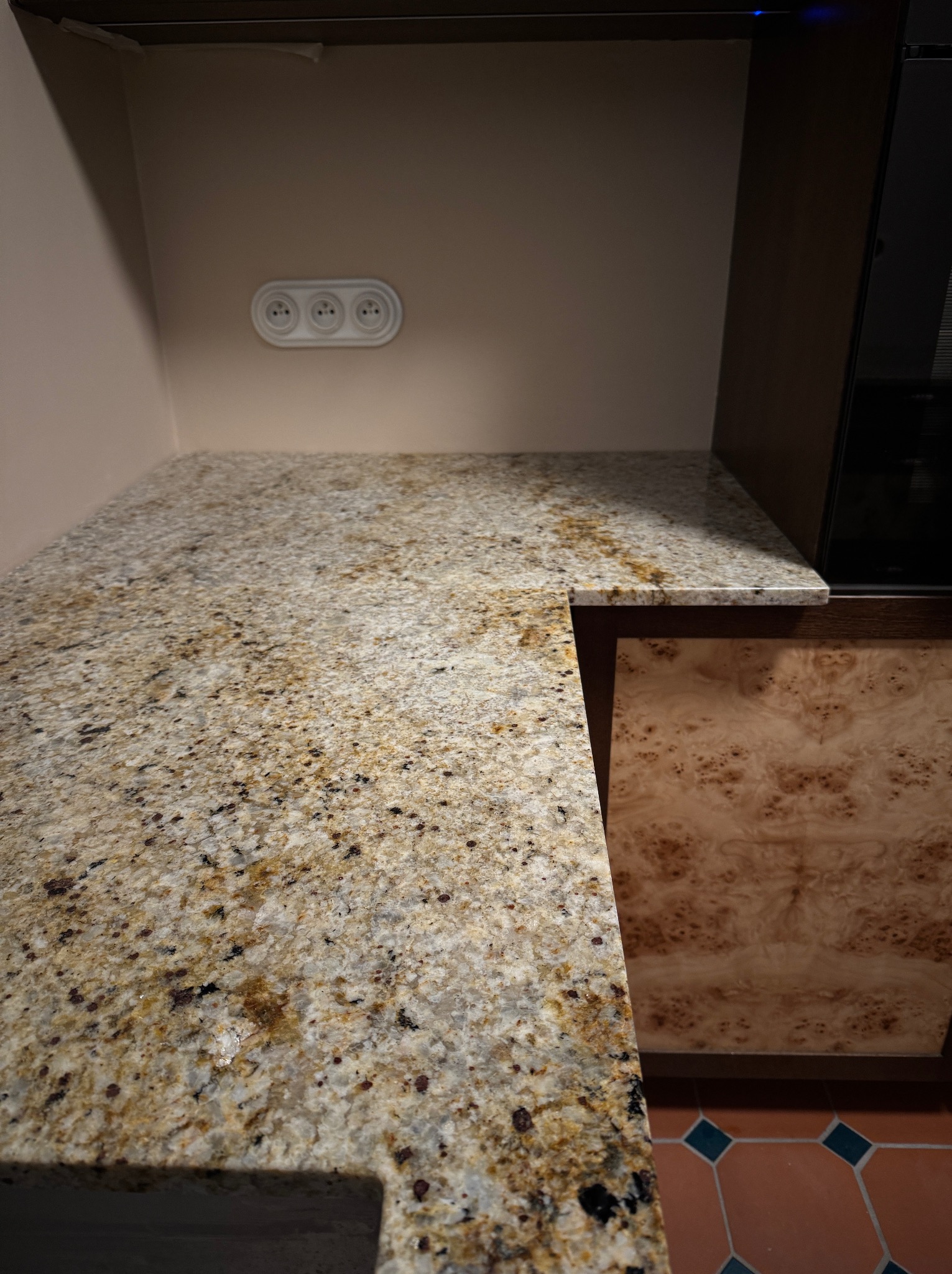 Beżowy blat kamienny z brązowymi i czarnymi plamkami, zamontowany w kuchni. Widoczne gniazdka elektryczne i fragment szafki z wzorzystym fornirem.