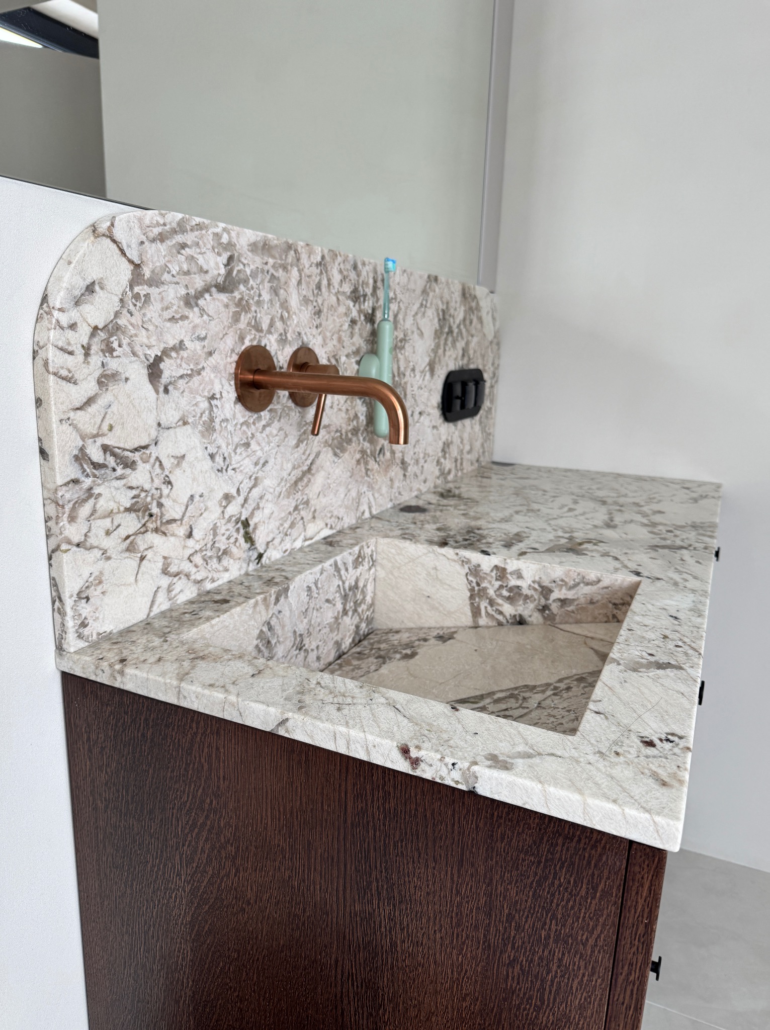 Biało-szary blat kamienny z wbudowaną umywalką i miedzianą armaturą na tle białej ściany. Umywalka z kamienia o wyraźnej fakturze, brązowa szafka pod umywalką.