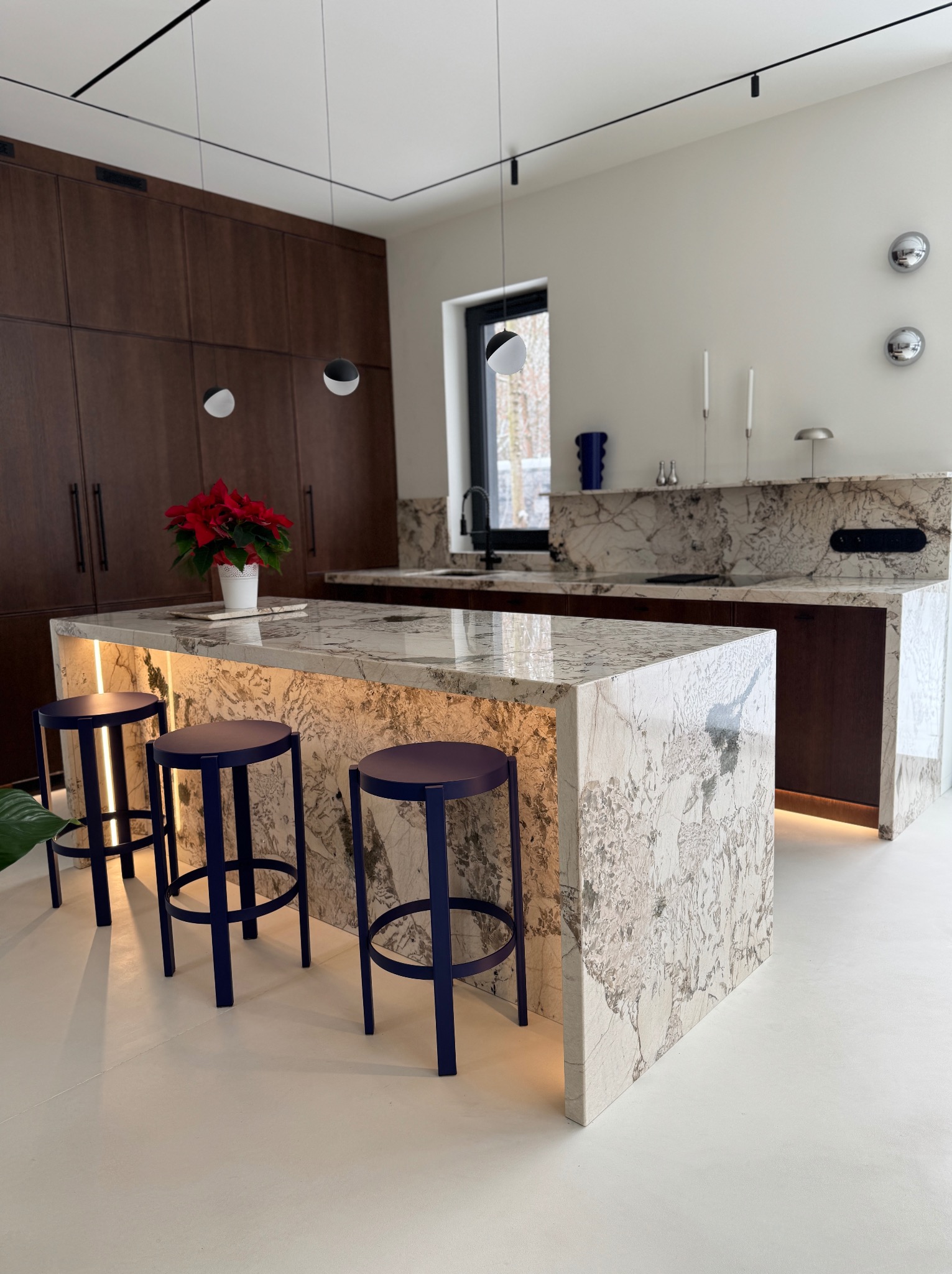 Nowoczesna kuchnia z wyspą i blatem z kamienia naturalnego o unikalnym wzorze, podświetlonym od spodu. Trzy niebieskie hokery przy wyspie, minimalistyczne oświetlenie.