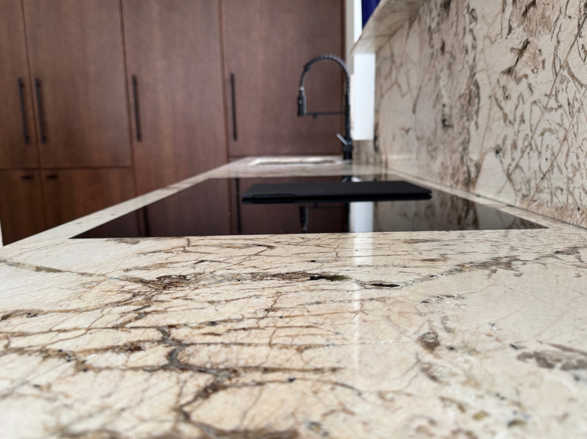 Beżowy blat kamienny z brązowymi żyłkami, na nim płyta indukcyjna. W tle szafki kuchenne i bateria. Ujęcie z niskiej perspektywy podkreśla wzór kamienia.