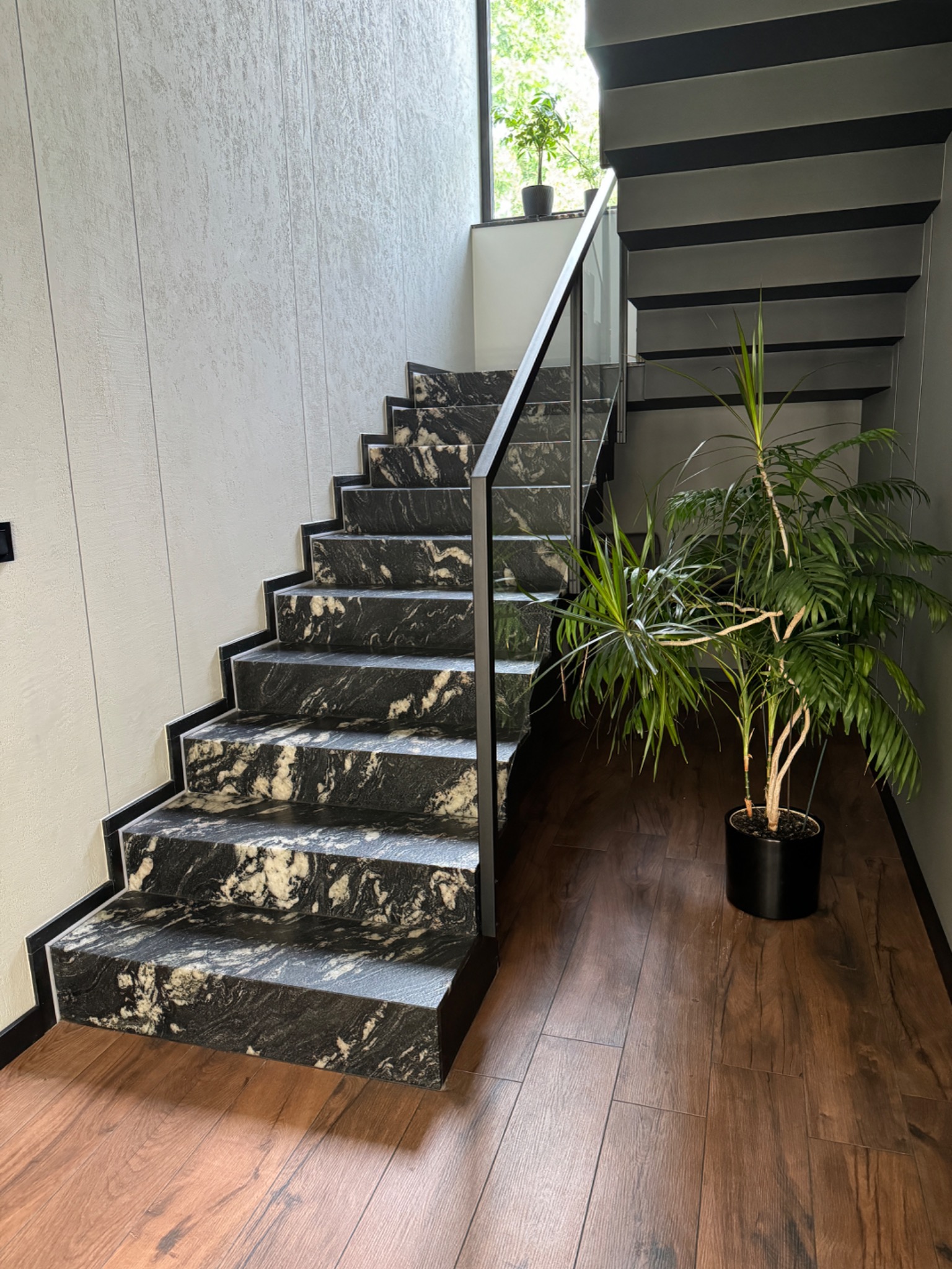 Nowoczesne schody z czarnego kamienia z białymi żyłami, z metalową balustradą i roślinami doniczkowymi, na drewnianej podłodze. Minimalistyczny design.