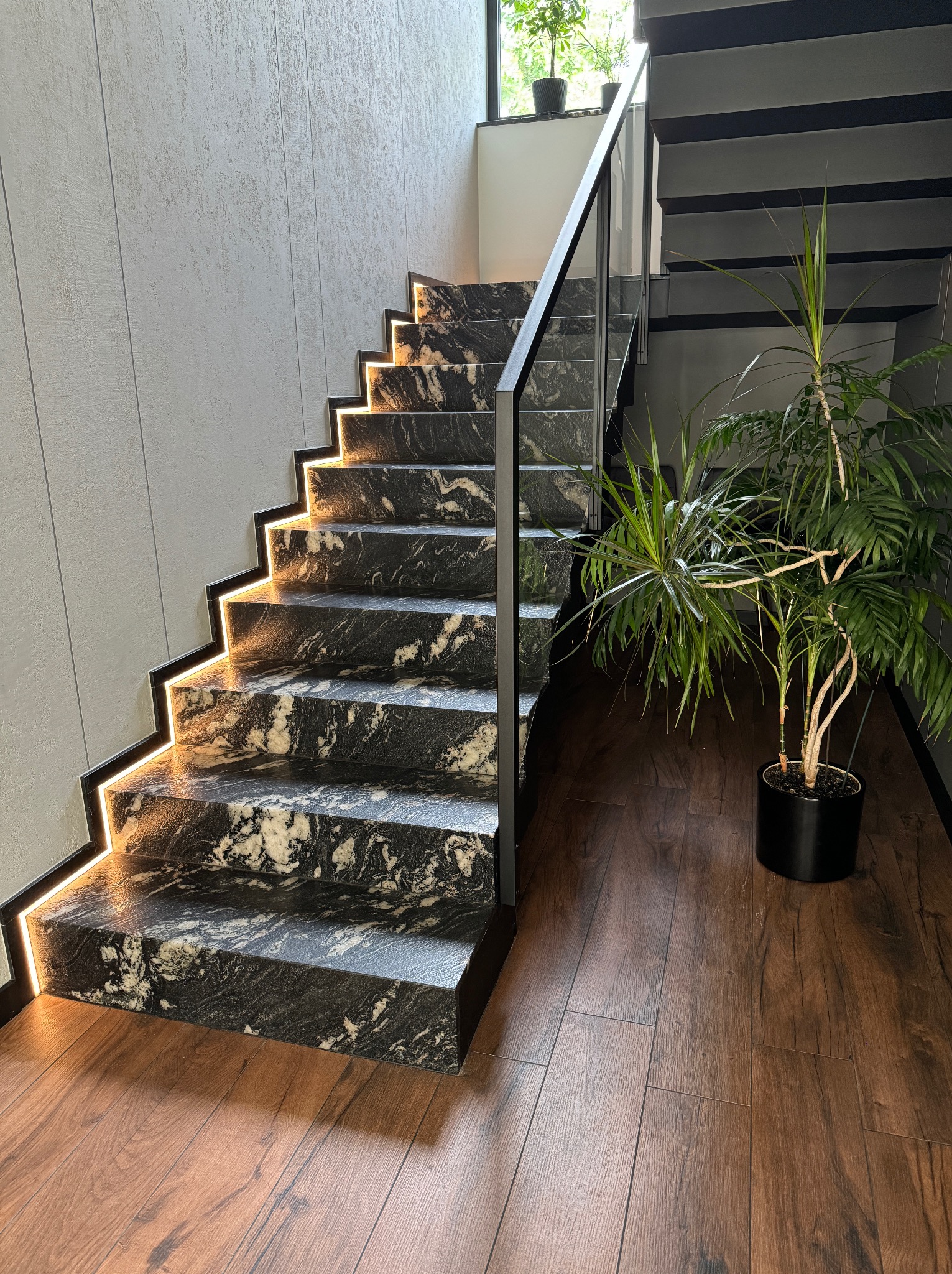 Schody z czarnego kamienia z białymi żyłami, podświetlone ciepłym światłem LED, z metalową balustradą. W tle widoczna zieleń roślin doniczkowych.