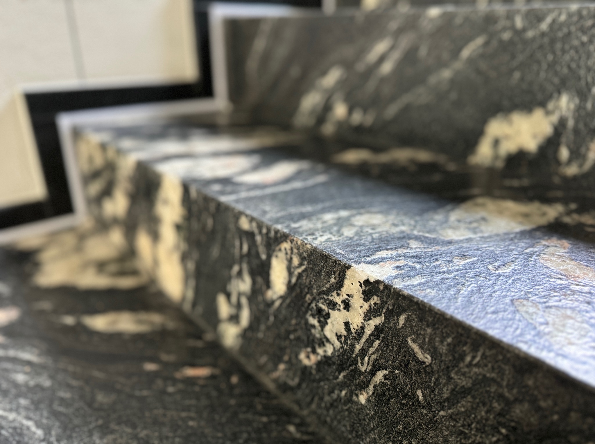 Schody z ciemnego kamienia z unikalnym wzorem, widoczne z bliska, z delikatnym rozmyciem tła. Krawędź stopnia oświetlona, podkreślająca fakturę kamienia.