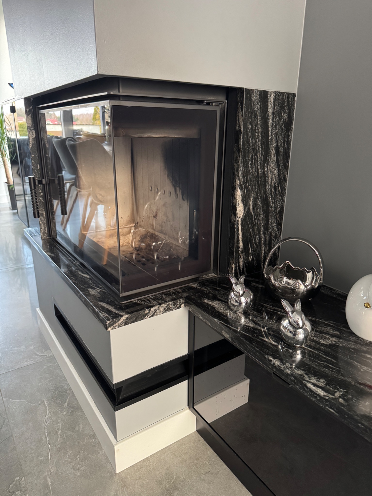 Nowoczesny kominek z blatem z czarnego kamienia o wyraźnym użyleniu, zdobiony srebrnymi figurkami zajączków i dekoracyjną misą. Szare tło i jasna podłoga.