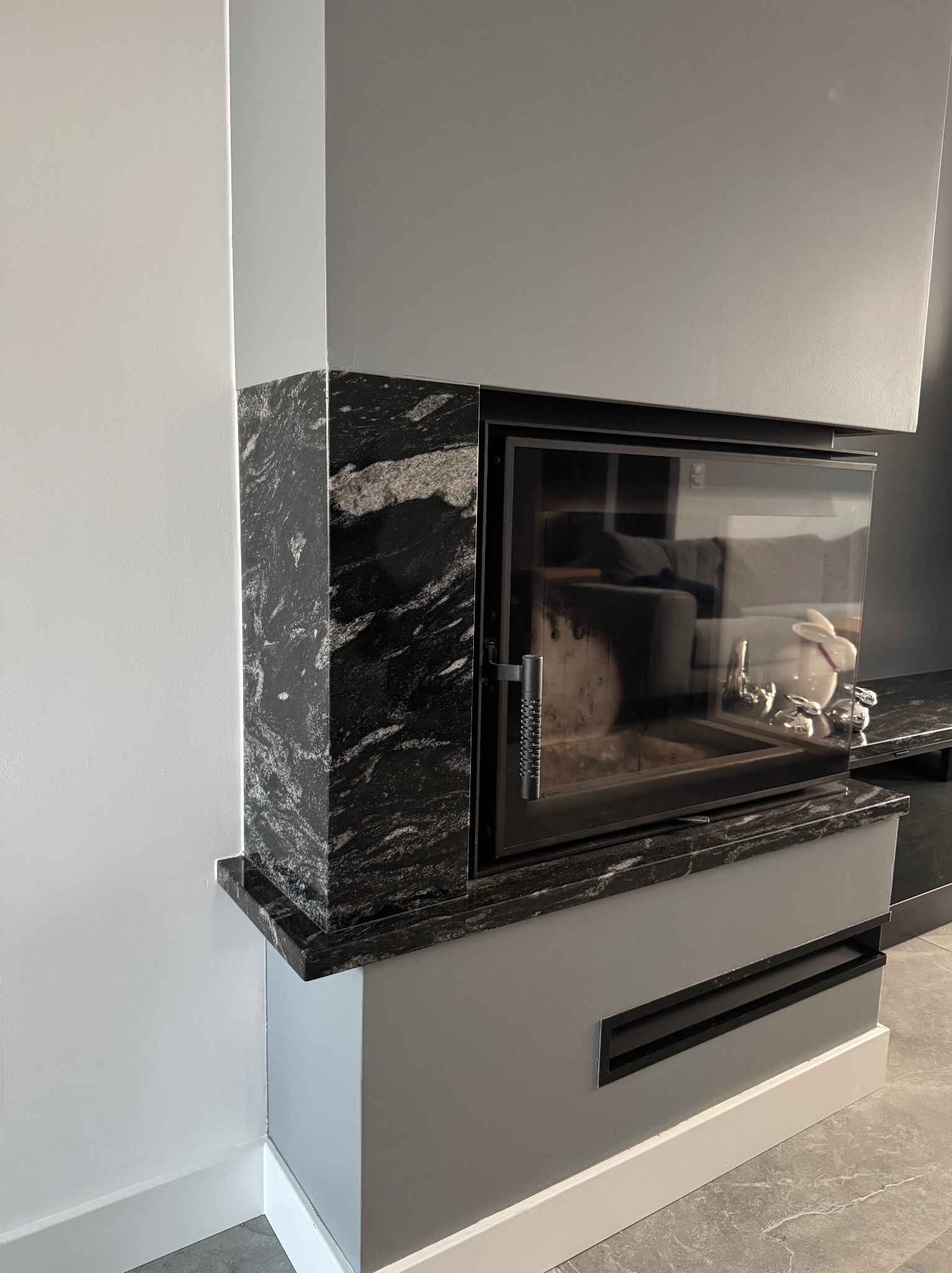 Nowoczesny kominek z obudową z czarnego, wzorzystego kamienia, z widocznym paleniskiem i elementami dekoracyjnymi wewnątrz. Szare ściany i podłoga.