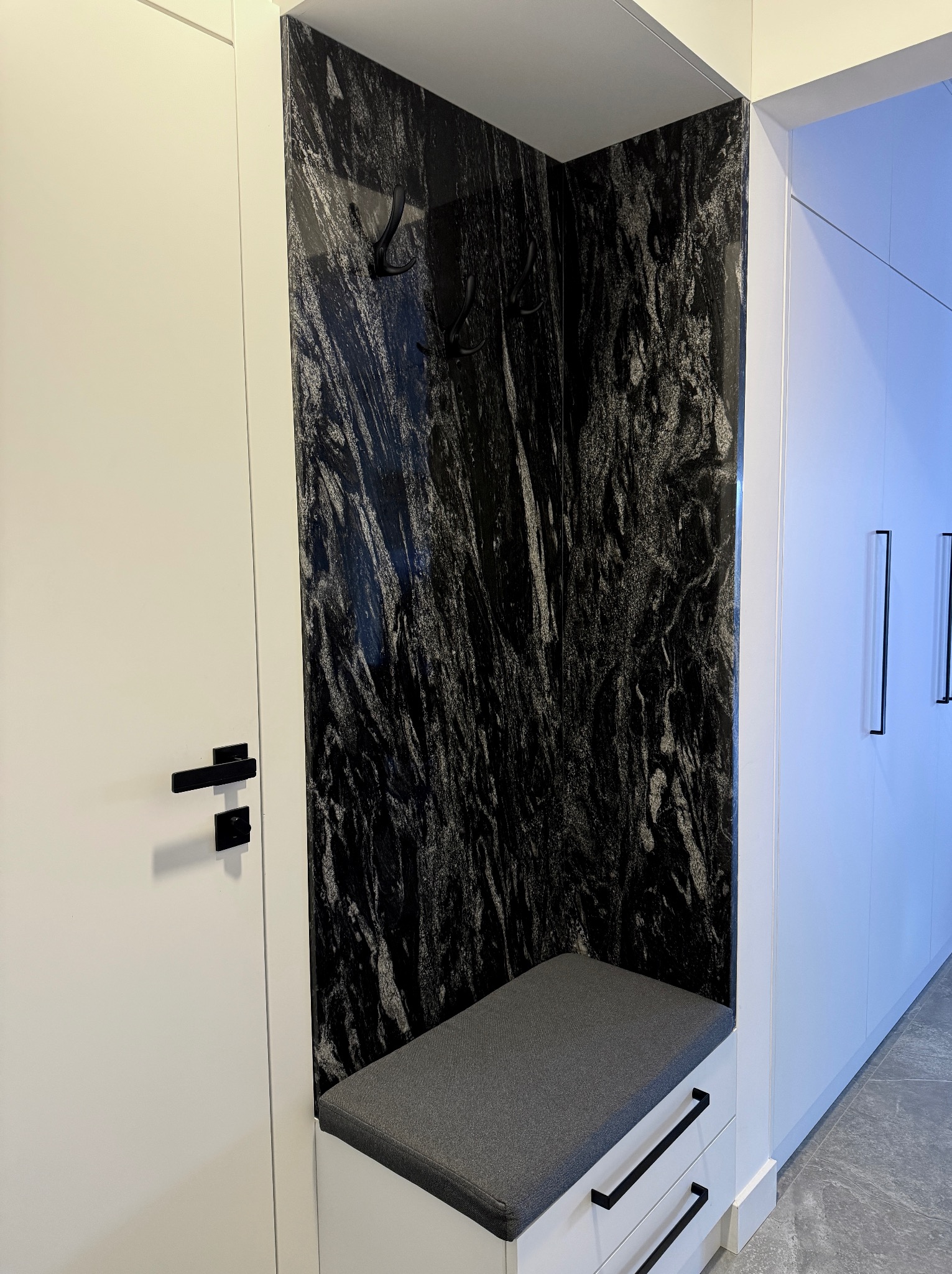 Wnęka z siedziskiem i czarnym, wzorzystym kamieniem na ścianie, uzupełniona o wieszaki. Minimalistyczny design, białe szafki i szuflady, czarne uchwyty.