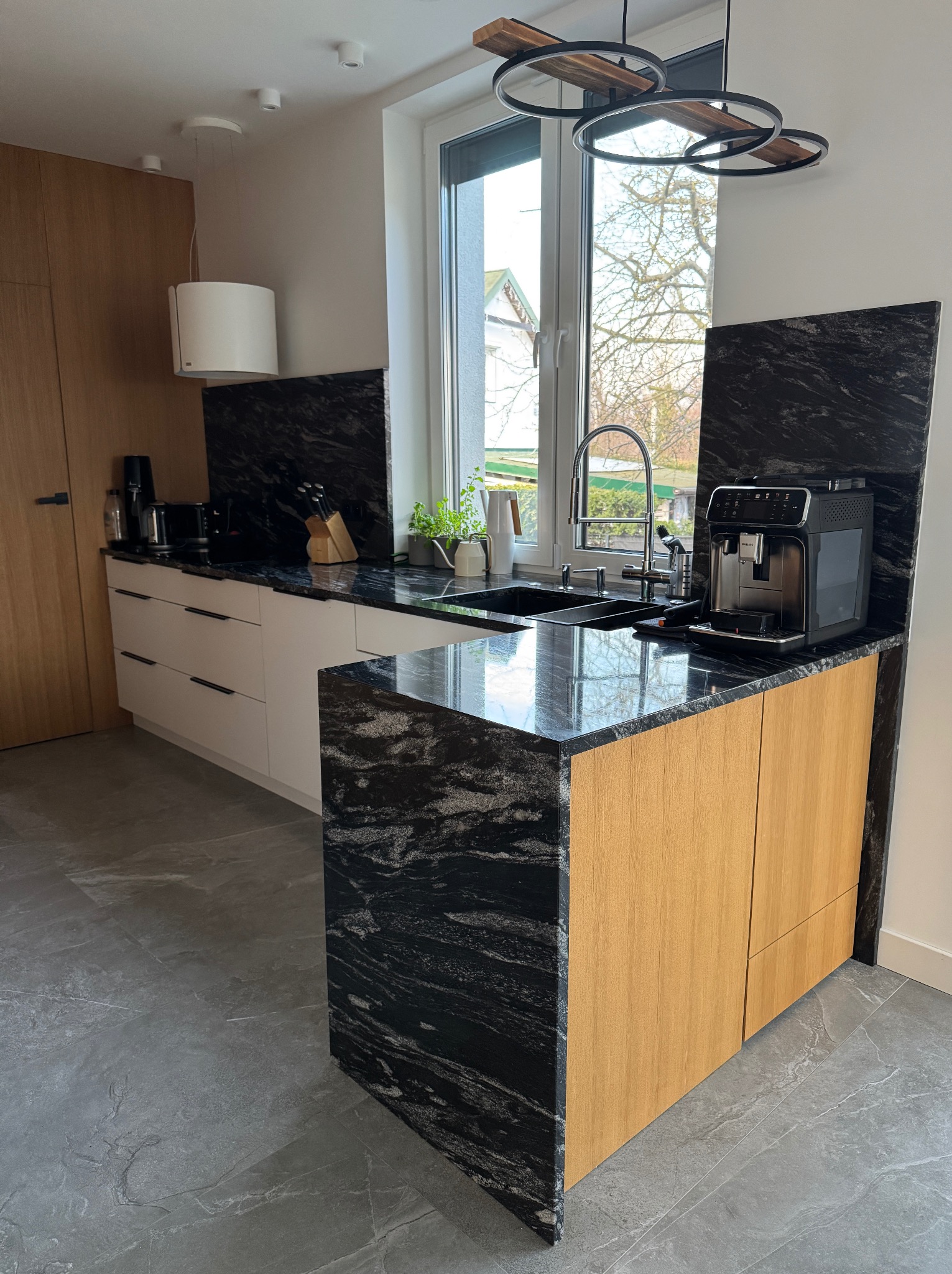 Kuchnia z blatem z czarnego kamienia o wyraźnym wzorze, drewniane szafki, nowoczesne oświetlenie i sprzęt AGD. Widok na okno z zielenią w tle.