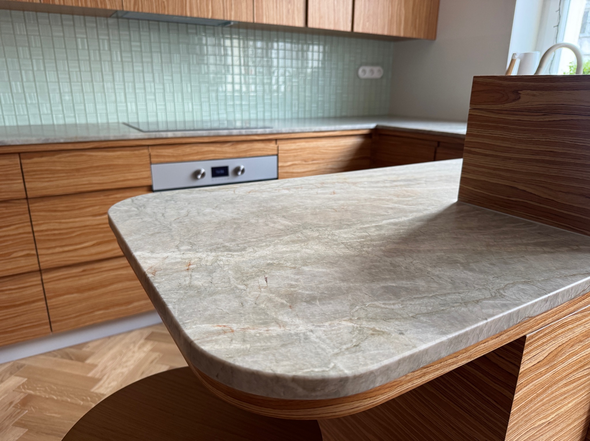 Marmurowy blat kuchenny z zaokrąglonymi krawędziami, drewniane szafki i mozaika na ścianie w tle. Nowoczesny design.