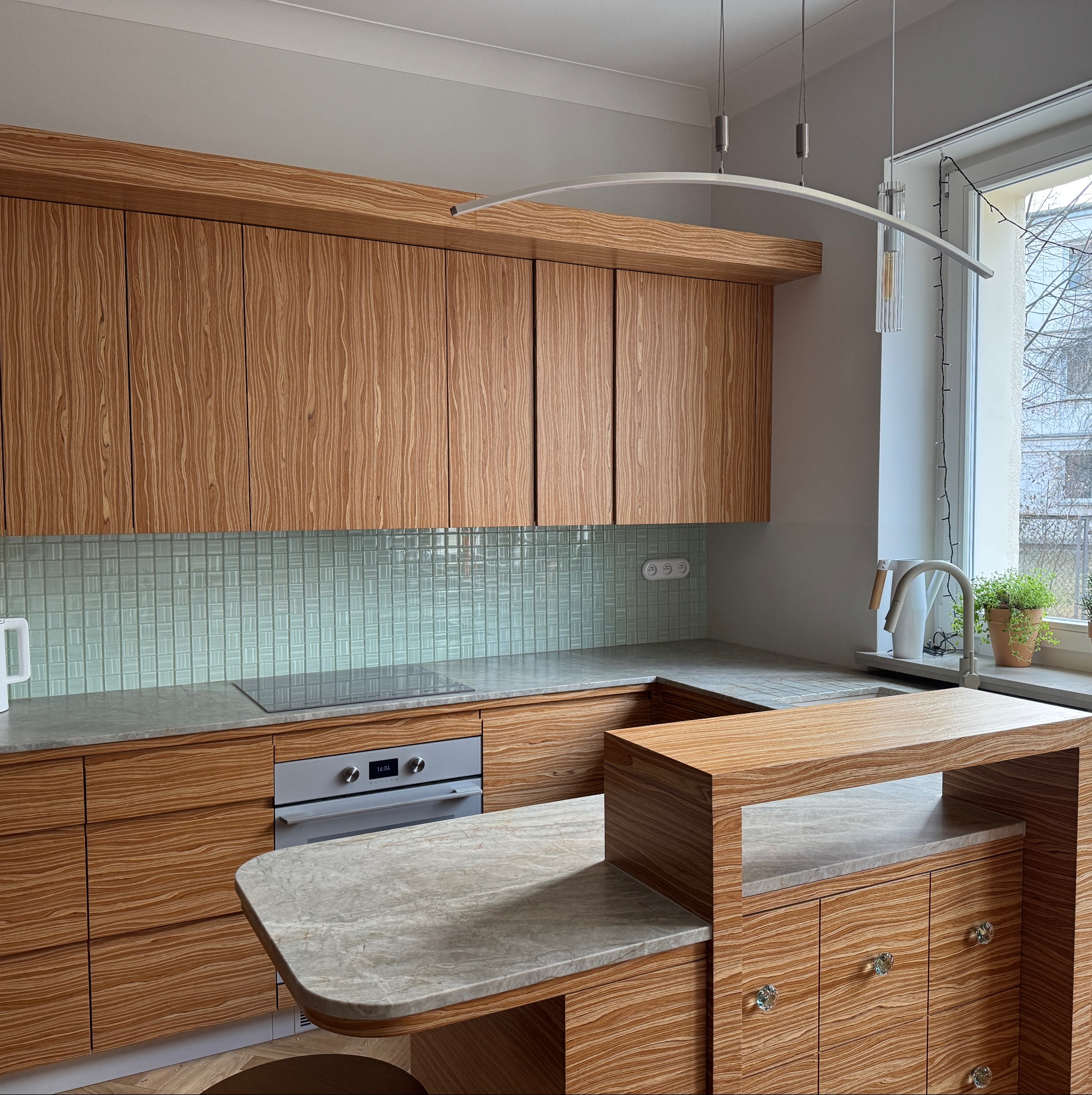 Nowoczesna kuchnia w Rudzienku z drewnianymi szafkami o unikalnym wzorze i blatem o marmurowej fakturze. Jasne wnętrze z roślinami w oknie.