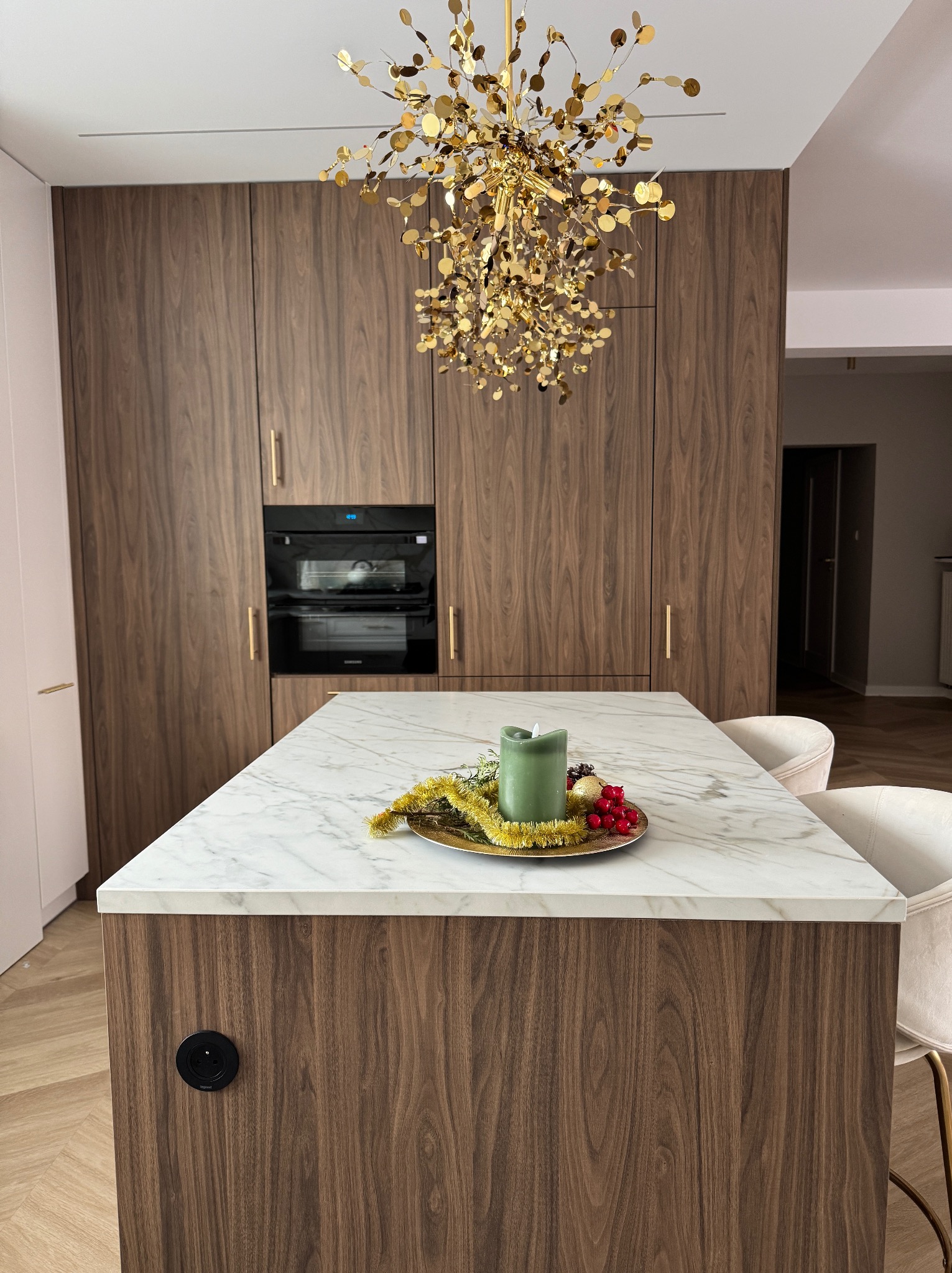 Marmurowy blat wyspy kuchennej z dekoracją świąteczną, na tle zabudowy z forniru orzechowego i złotego żyrandola. Nowoczesne wnętrze kuchni.