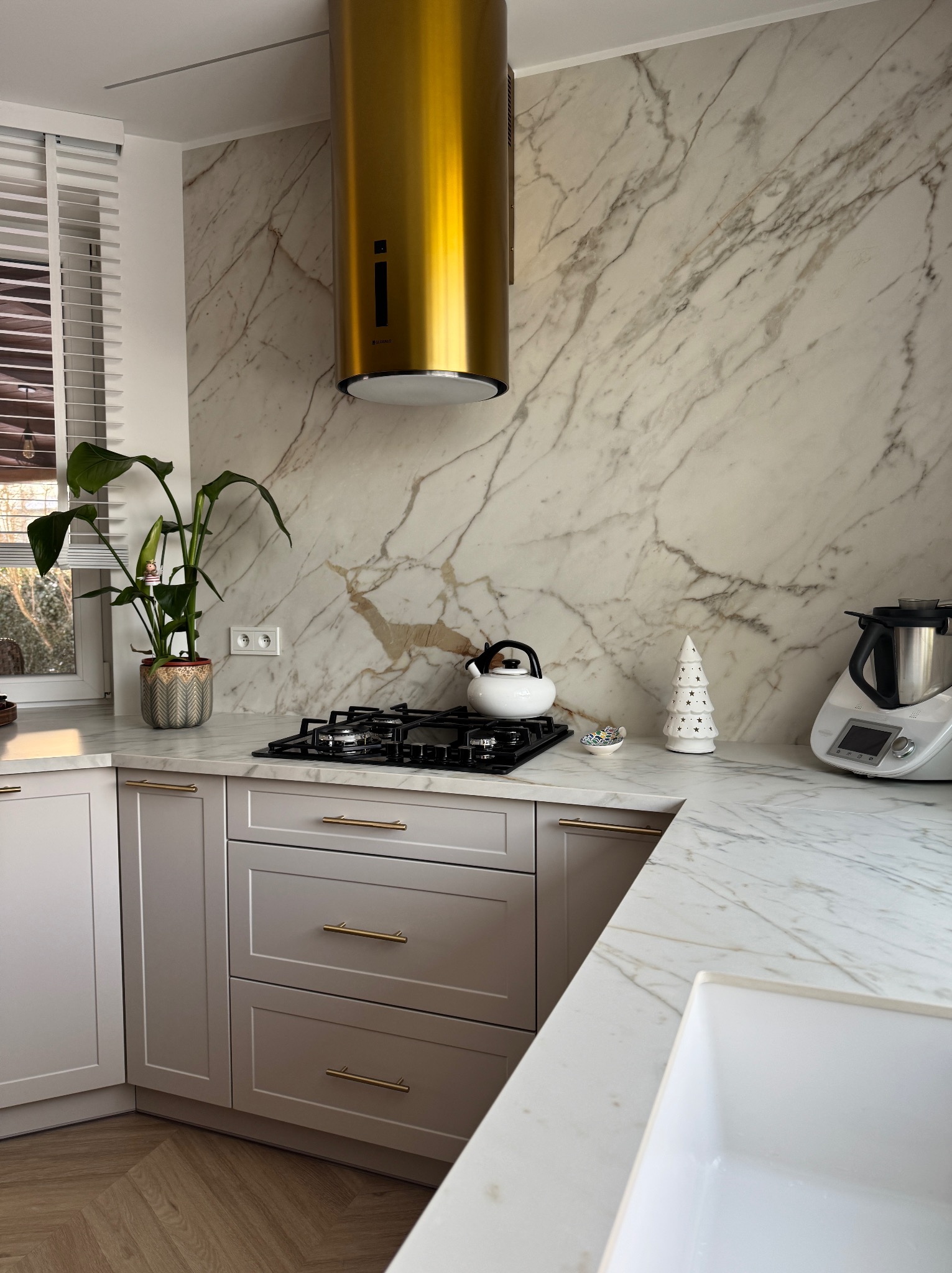 Kuchnia z blatem z kamienia imitującego marmur, złoty okap, szare szafki z złotymi uchwytami, roślina w doniczce i biały czajnik na płycie grzewczej.