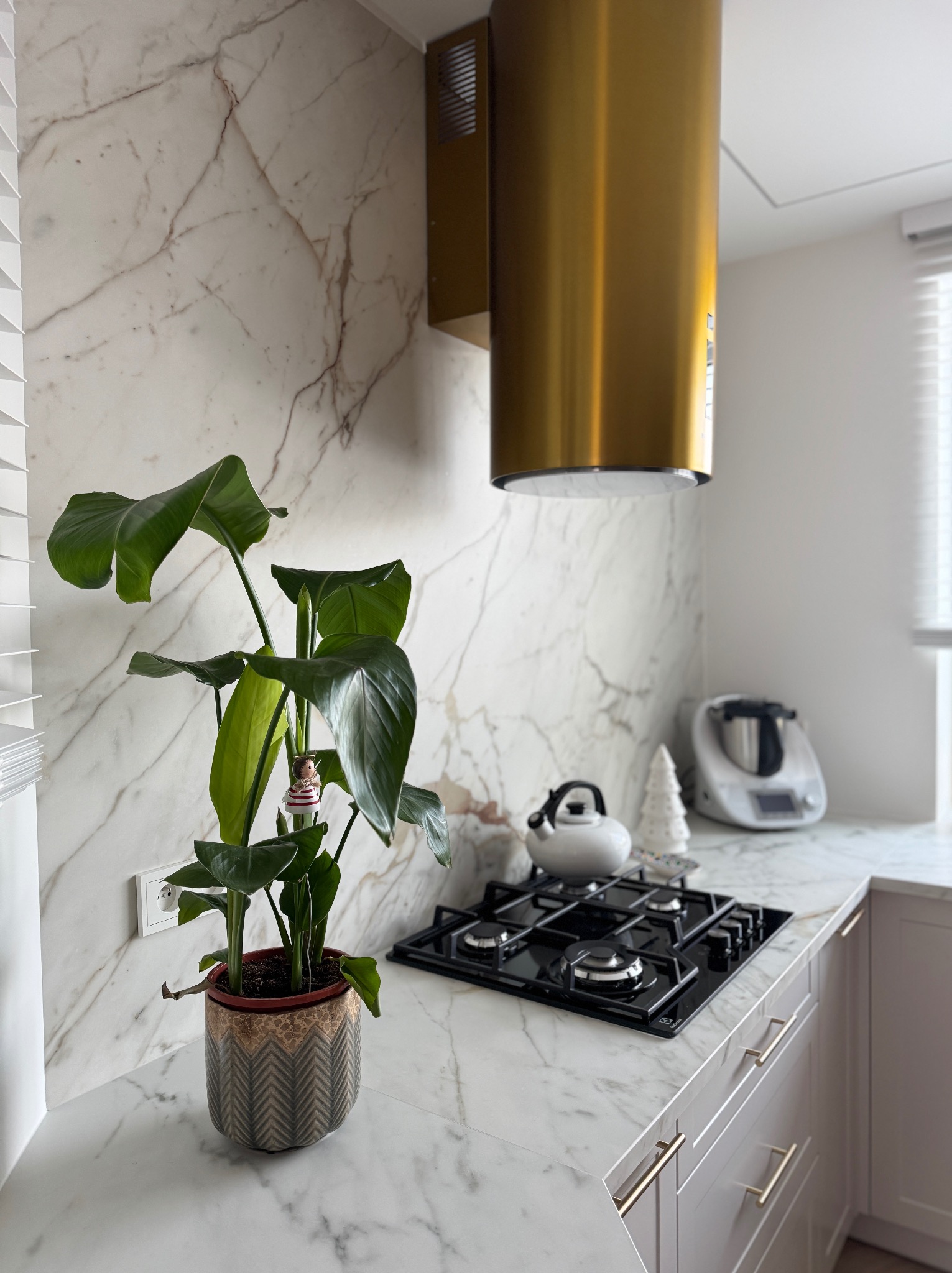 Biały blat kuchenny z marmurowym wzorem, czarna płyta grzewcza, złoty okap i roślina doniczkowa. Nowoczesna kuchnia z elementami dekoracyjnymi.