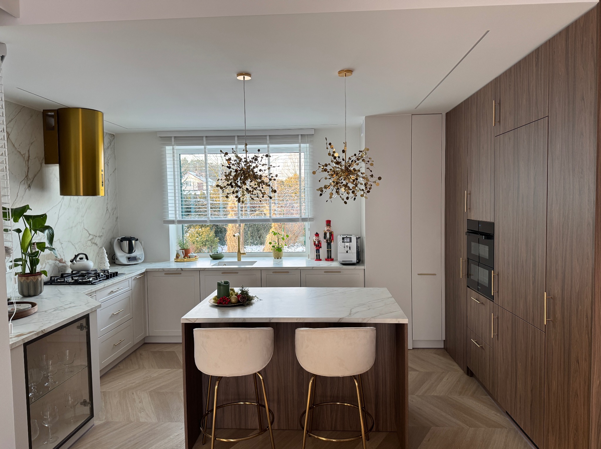 Elegancka kuchnia z wyspą i blatem z białego kamienia. Złote detale, nowoczesne oświetlenie i parkiet w jodełkę tworzą luksusową przestrzeń. Widok na zewnątrz przez okno.