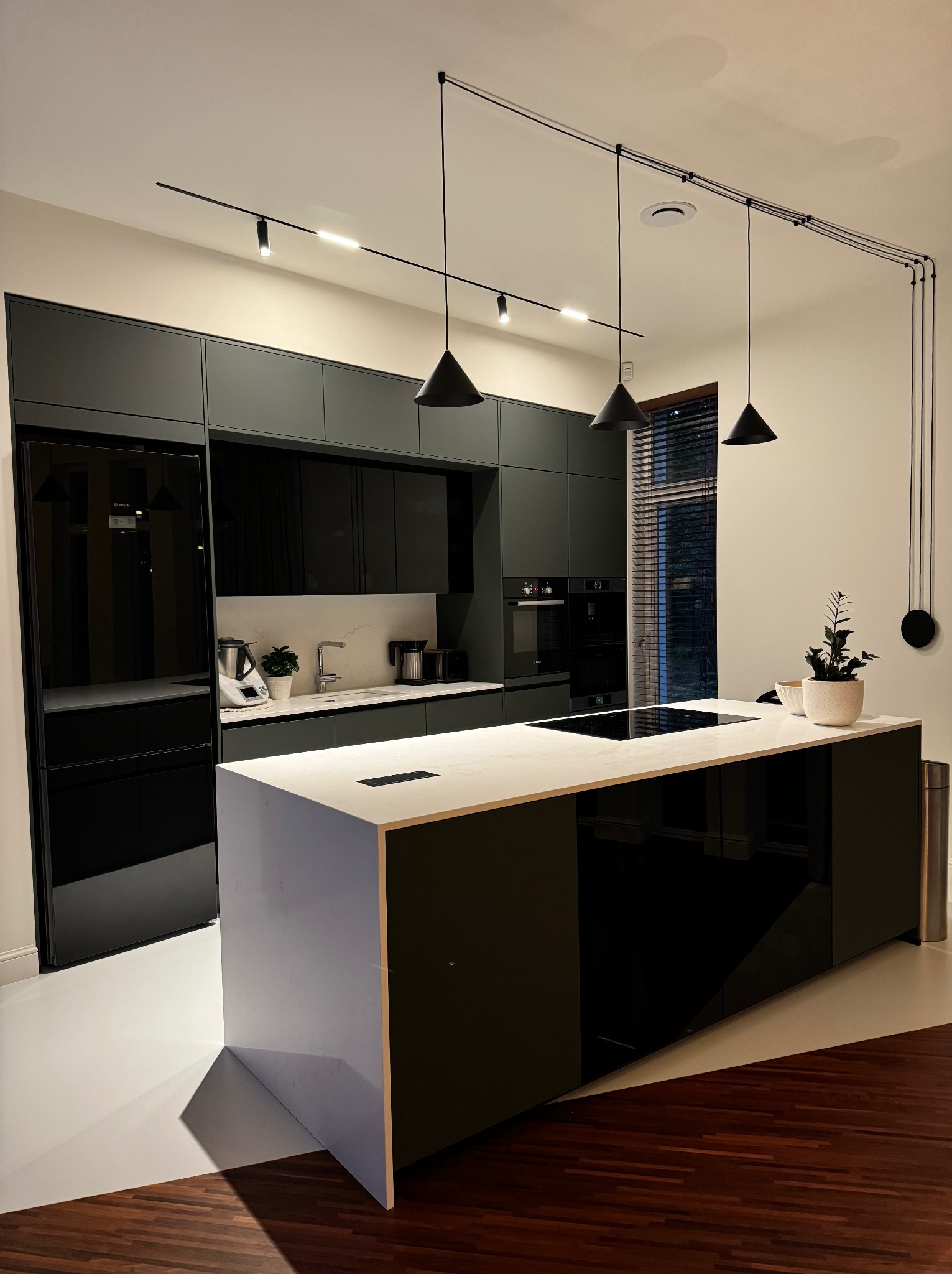 Nowoczesna kuchnia z wyspą i blatem w kolorze bieli, czarne szafki, oświetlenie szynowe. Minimalistyczny design, drewniana podłoga, roślina w doniczce.