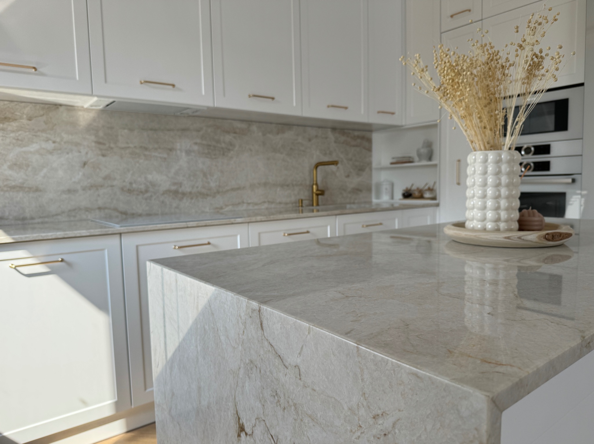 Jasny blat kamienny z subtelnym użyleniem w nowoczesnej kuchni z białymi szafkami i złotą armaturą. Na blacie dekoracyjny wazon z suszem.