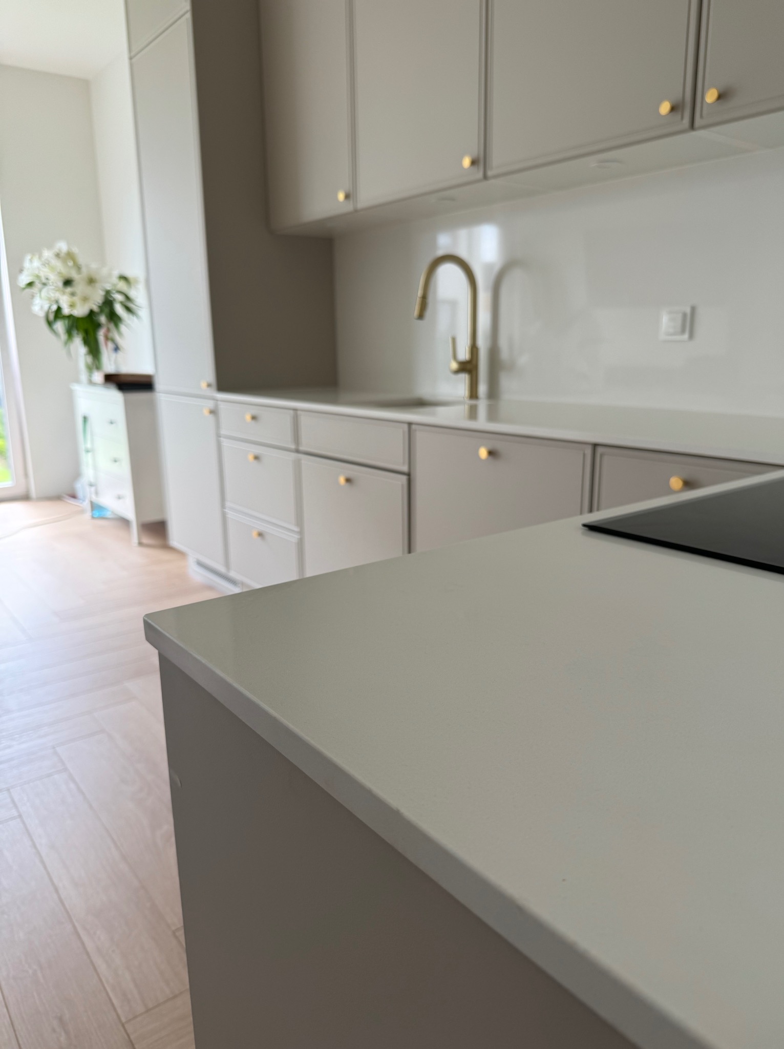 Jasny blat kuchenny z płytą indukcyjną, złota bateria i kremowe szafki z okrągłymi uchwytami. W tle kwiaty i parkiet w jodełkę.