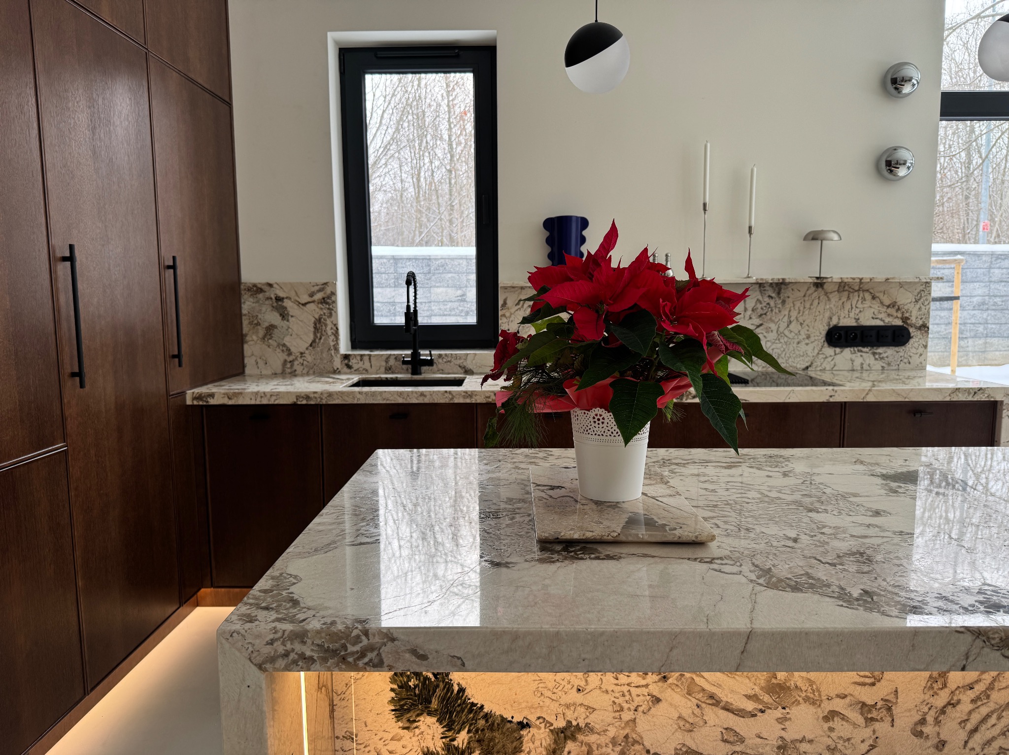 Kuchnia z wyspą i blatem z jasnego kamienia o wyraźnym wzorze. Na blacie stoi biała doniczka z czerwoną gwiazdą betlejemską. W tle okno i zabudowa kuchenna.