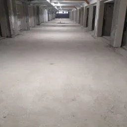 Długi korytarz w surowym stanie, przygotowany pod wylewkę, widoczne liczne kolumny i drzwi po bokach, oświetlenie sufitowe.