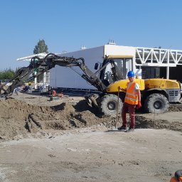 GAJOWSKI SEBASTIAN, KMh - Operator koparki kołowej Volvo 12 MTX w pomarańczowej kamizelce i niebieskim kasku, trzymający łopatę na placu budowy z wykopanym terenem, w tle widoczny szkielet budynku.