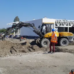 Operator koparki kołowej Volvo 12 MTX w pomarańczowej kamizelce i niebieskim kasku, trzymający łopatę na placu budowy z wykopanym terenem, w tle widoczny szkielet budynku.