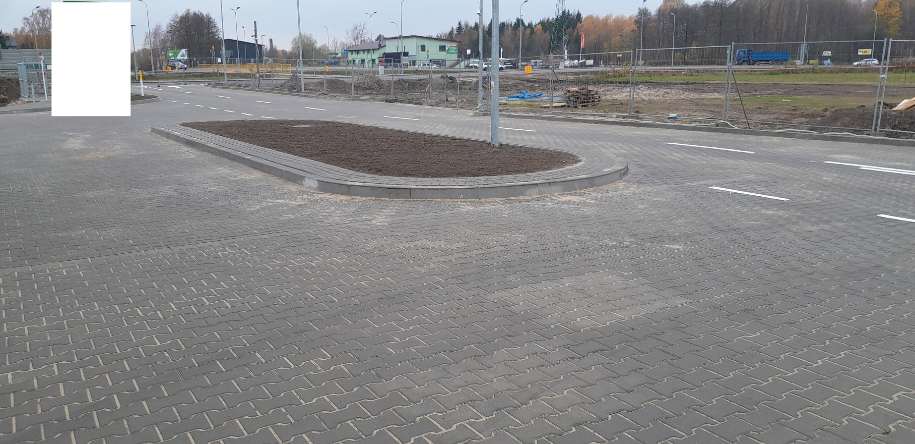 Nowo ułożona szara kostka brukowa w jodełkę na terenie przemysłowym z wysepką obsadzoną ziemią, widoczne białe pasy na jezdni.