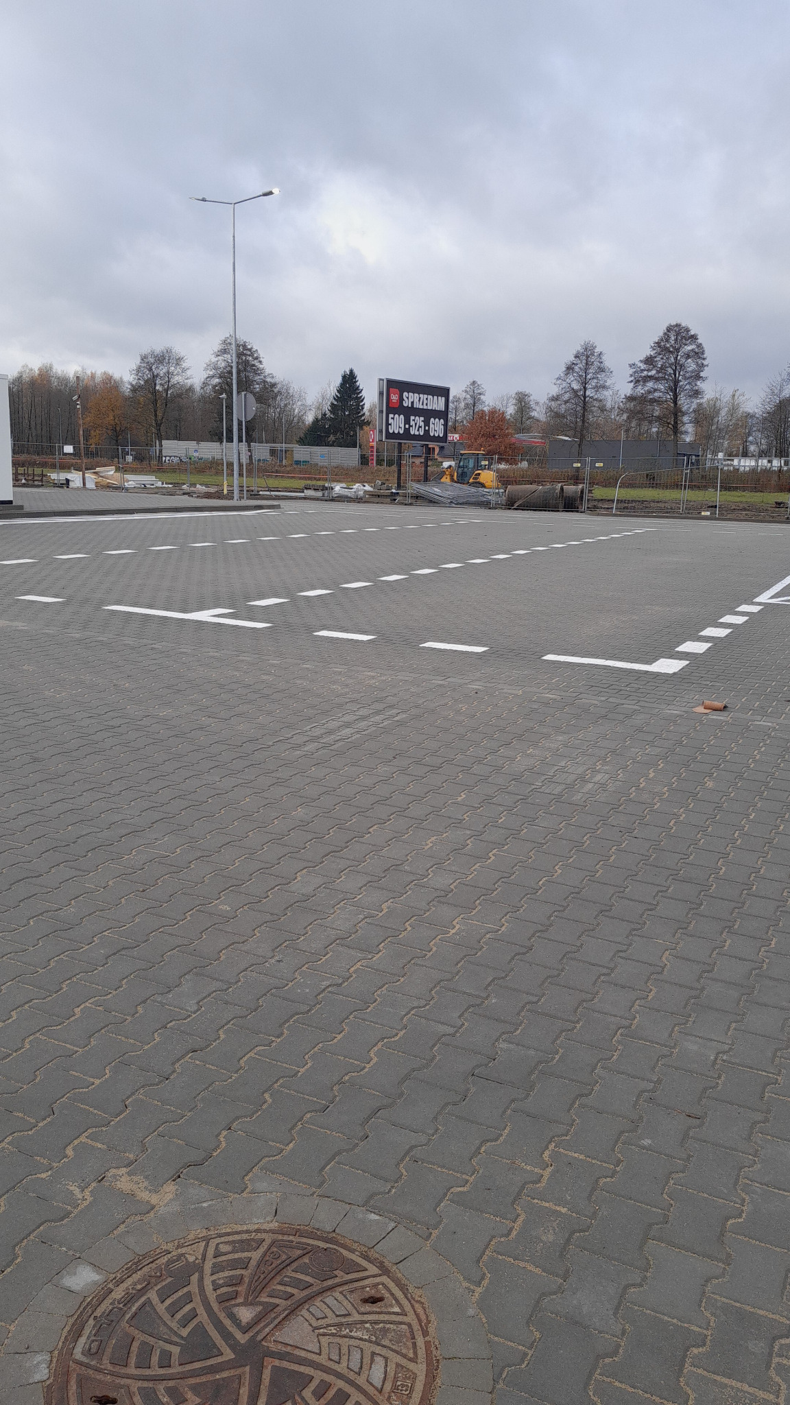 Nowo ułożona szara kostka brukowa z białymi liniami wyznaczającymi miejsca parkingowe, w tle widoczny billboard reklamowy i maszyna budowlana.