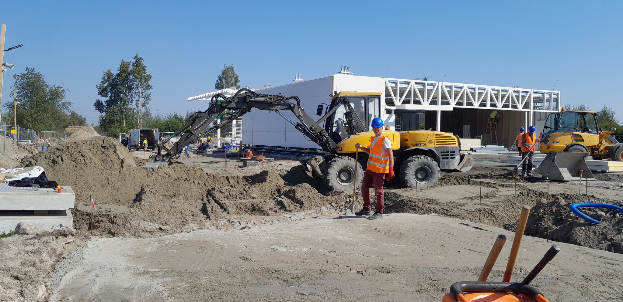 Operator koparki kołowej Volvo 12 MTX w pomarańczowej kamizelce i niebieskim kasku, trzymający łopatę na placu budowy z wykopanym terenem, w tle widoczny szkielet budynku.