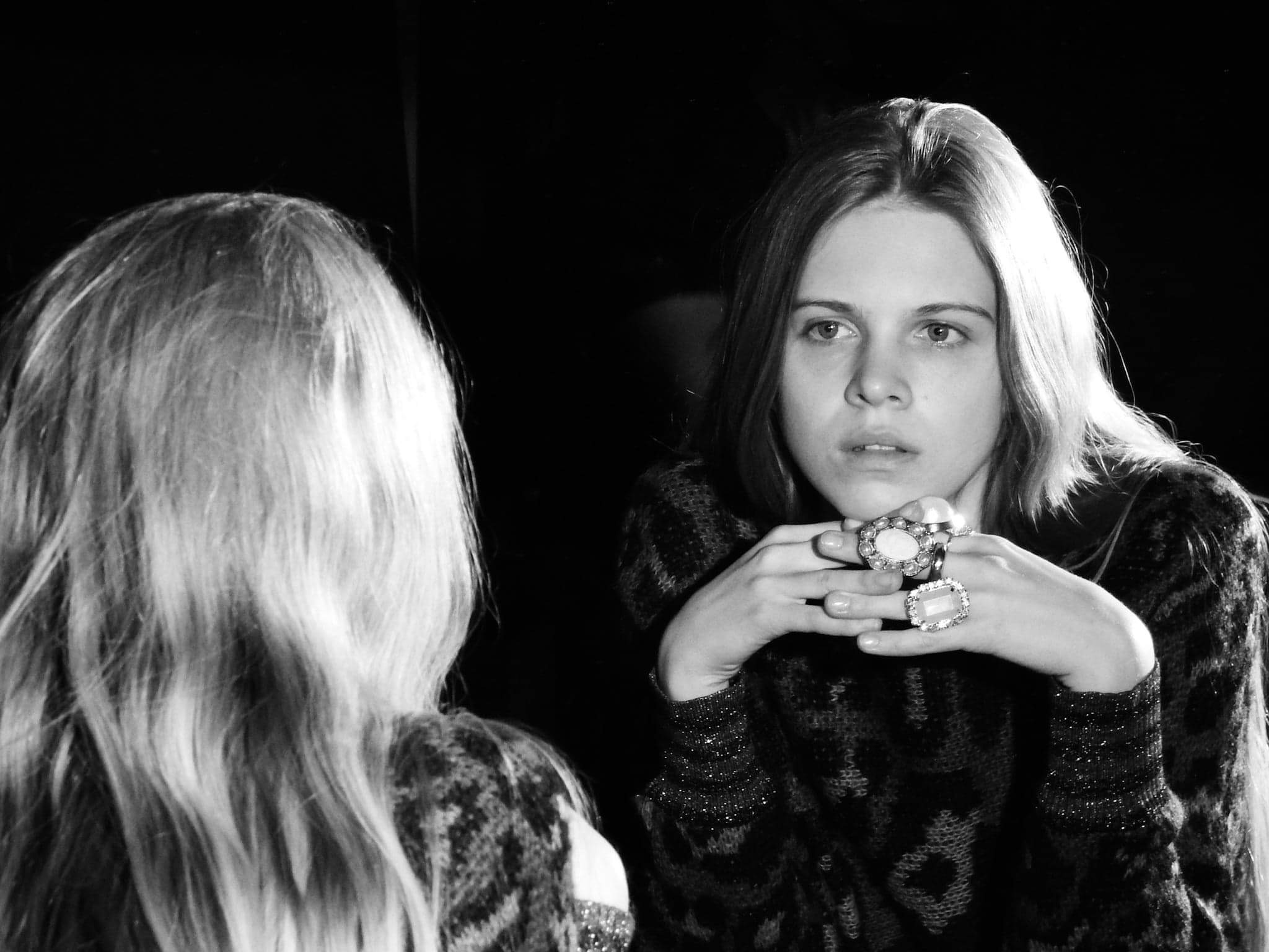 Czarno-biały portret kobiety z dwoma dużymi pierścionkami na dłoniach, odbicie w lustrze z widocznymi włosami i częścią ramienia.