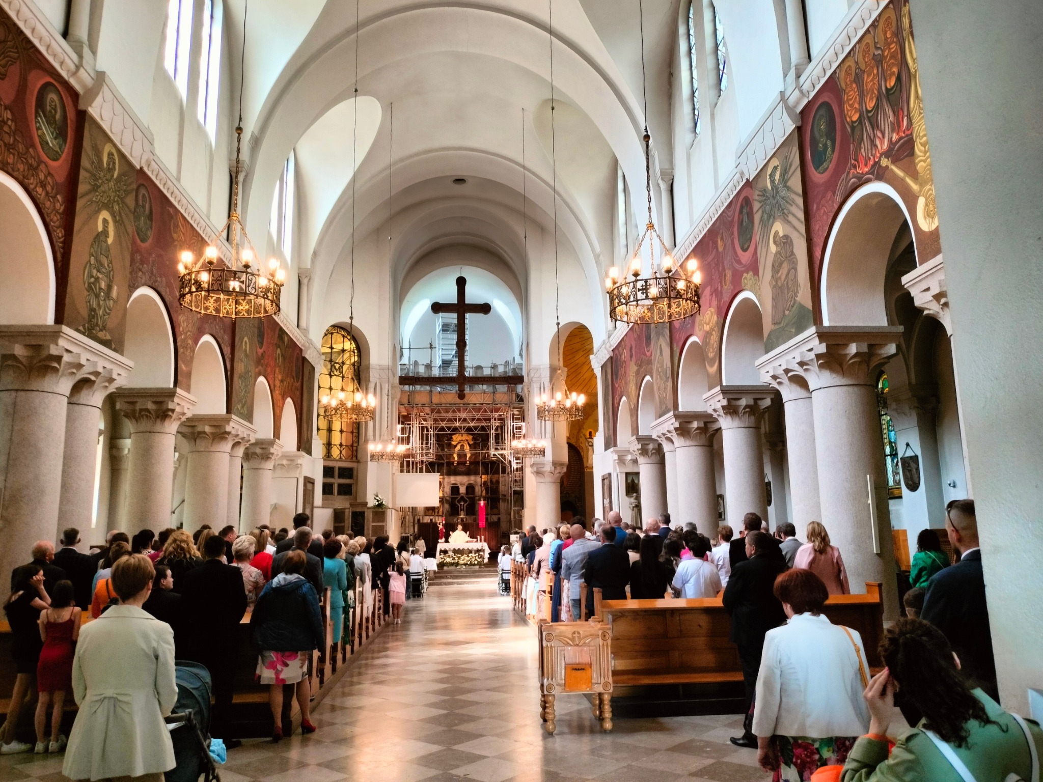 Wnętrze kościoła podczas uroczystości Pierwszej Komunii Świętej, widok na ołtarz przez nawę główną, uczestnicy zgromadzeni w ławkach, dekoracje kwiatowe i oświetlenie.