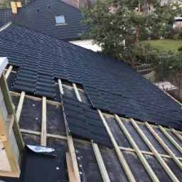 Montaż czarnej blachodachówki na drewnianej konstrukcji dachu, widoczne elementy mocujące i membrana dachowa, w tle sosna i zabudowa jednorodzinna.