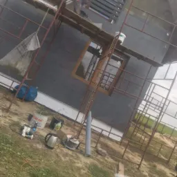 Prace na elewacji domu jednorodzinnego w trakcie realizacji, widoczne rusztowania, ocieplenie w kolorze grafitowym, okna z drewnianymi ramami, pracownik na rusztowaniu w kombinezonie i masce...