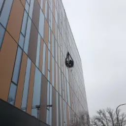 Wysoki budynek biurowy z elewacją w odcieniach brązu i szarości, na wysokości zawieszony kosz z pracownikami, widok z dołu na pochmurne niebo.