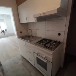 Nowoczesna biała kuchnia z płytą gazową i piekarnikiem, widok na sąsiednie pomieszczenie w trakcie prac wykończeniowych, okno z torbą roboczą.