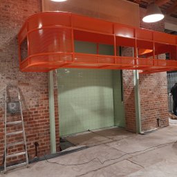 K&K BROTHERS RENOVATION - Wnętrze w trakcie remontu z pomarańczową, ażurową konstrukcją pod sufitem, widoczna zielona ściana z płytek, mur z czerwonej cegły, drabina i pracownicy z odkurzaczem przemysłowym.