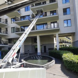 K&K BROTHERS RENOVATION - Wysięgnik dźwigu meblowego CEZAR LIFT z numerem telefonu 0477/315.651 ustawiony przed nowoczesnym budynkiem apartamentowym z balkonami, fontanna w tle.