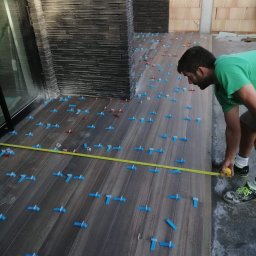 K&K BROTHERS RENOVATION - Mężczyzna w zielonej koszulce mierzy taśmą ułożone brązowe płytki podłogowe z niebieskimi i czerwonymi klipsami poziomującymi, widoczna fragment ściany z kamienia dekoracyjnego.