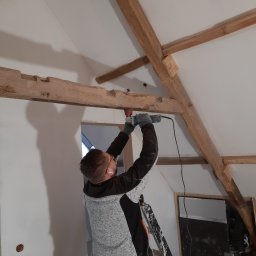 K&K BROTHERS RENOVATION - Pracownik szlifuje drewnianą belkę stropową na poddaszu za pomocą szlifierki kątowej, widoczna drabina i instalacja elektryczna w ścianie.