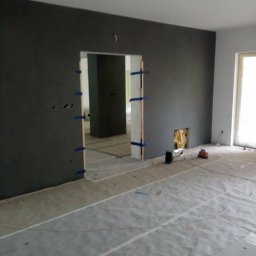 K&K BROTHERS RENOVATION - Pokój w trakcie malowania: ściany w ciemnym odcieniu szarości, zabezpieczona podłoga, otwór drzwiowy z listwami przyklejonymi taśmą malarską, widoczne narzędzia malarskie.