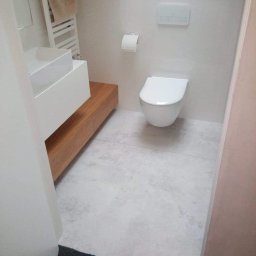 K&K BROTHERS RENOVATION - Nowoczesna łazienka z wiszącą toaletą, umywalką nablatową na drewnianej szafce i szarą, betonową posadzką, widok zza futryny drzwi.