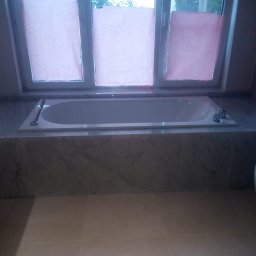 K&K BROTHERS RENOVATION - Wanna w zabudowie z marmurową obudową, z baterią i chromowaną słuchawką, pod oknem z różowymi zasłonami.