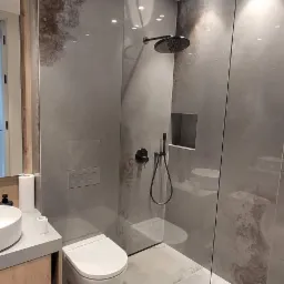 Nowoczesna łazienka z kabiną prysznicową walk-in, szare płytki imitujące beton, wisząca toaleta i umywalka nablatowa na drewnianej szafce.